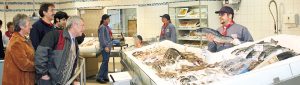 Zu sehen ist eine Aufnahme der Fischabteilung. Ein Mitarbeiter steht rechts hinter der Theke und präsentiert interessierten Kunden einen länglichen Fisch. Die Fischtheke vor ihm ist mit Eis und frischem Fisch befüllt. Im Hintergrund sind weitere Kunden, sowie drei weitere Mitarbeiter zu erkennen, die sich miteinander unterhalten.