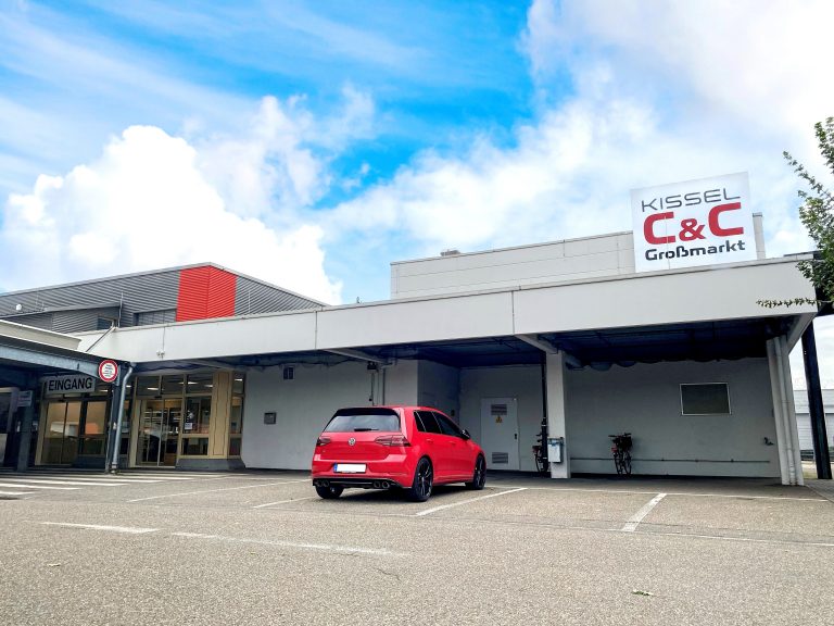 Zu sehen ist eine Außenaufnahme des Kissel C&C Großmarkt Landau. Rechts am Markt befestigt ist ein großes Schild mit dem Kissel C&C Großmarkt Logo. Links im Bild zu erkennen der Eingang des Marktes. Auf dem Parkplatz vor dem Gebäude steht ein roter VW.