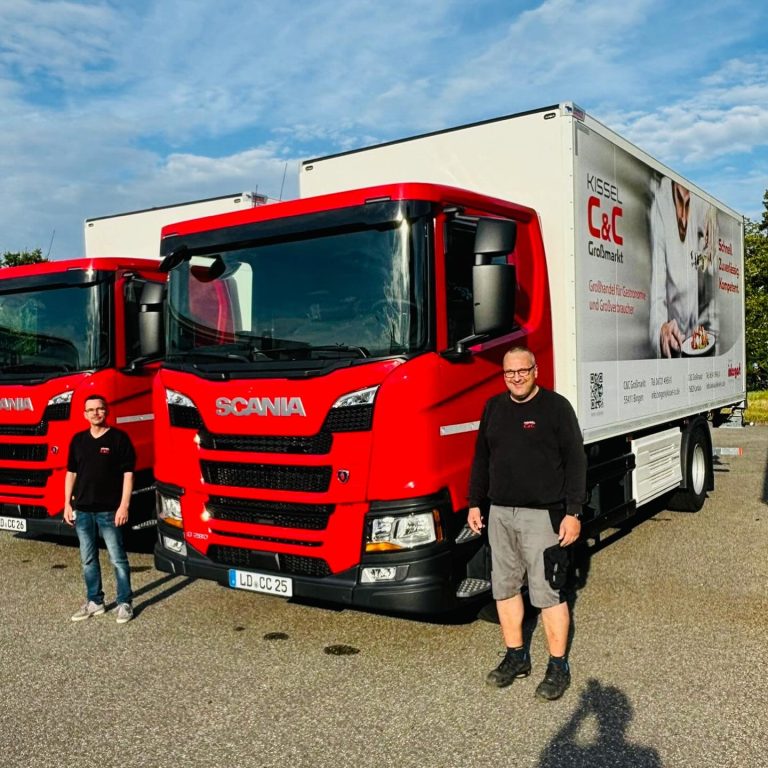 Zu sehen ist eine Fotografie von zwei C&C Mitarbeitern, die neben zwei neuen Ergänzungen des Fuhrparks stehen. Die neuen LKWs haben ein rotes Führerhaus, der Anhänger ist bedruckt mit dem Logo von "Kissel C&C Großmarkt".