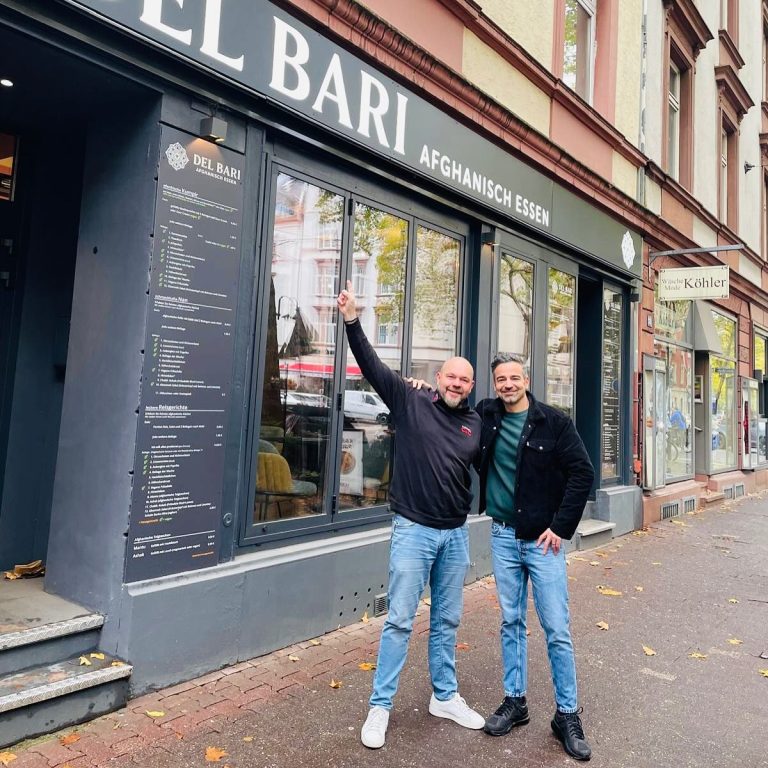 Zu sehen ist eine Aufnahme von einem C&C-Außendienstmitarbeiter zusammen mit einem Kunden, vor dem Restaurant "Del Bari" in Frankfurt. Der Mitarbeiter hat seinen rechten Arm in die Luft gestreckt, der Restaurantbesitzer und Kunde hat seine Hand lässig an der Hüfte liegen. Beide schauen lächelnd in die Kamera. Im Hintergrund ist der Restauranteingang des afghanischen Restaurants zu sehen. Eine Menükarte hängt vor dem Eingang aus.