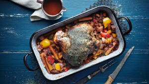 Man sieht eine geschmorte Lammkeule in einer rechteckigen Bratform. Die Lammkeule liegt mittig und ist von einer dunklen, würzigen Kräuterkruste bedeckt, die ihr eine rustikale und aromatische Optik verleiht. Rundherum befinden sich grob geschnittenes Gemüse wie gelbe und rote Paprikastücke, Karotten, Zwiebeln und Tomaten, die im Bratensaft weich geschmort sind. Die Zutaten wirken saftig und glänzend, was auf eine lange Garzeit schließen lässt. Die Bratform steht auf einem blau gestrichenen Holztisch, der einen schönen Kontrast zu den warmen Farben des Gerichts bildet. Daneben liegen ein Messer und eine Fleischgabel, während im oberen Bereich eine kleine Schale mit dunkler Sauce platziert ist. Insgesamt vermittelt das Bild den Eindruck eines herzhaften, langsam zubereiteten Ofengerichts mit kräftigen Aromen und traditionellem Charakter.